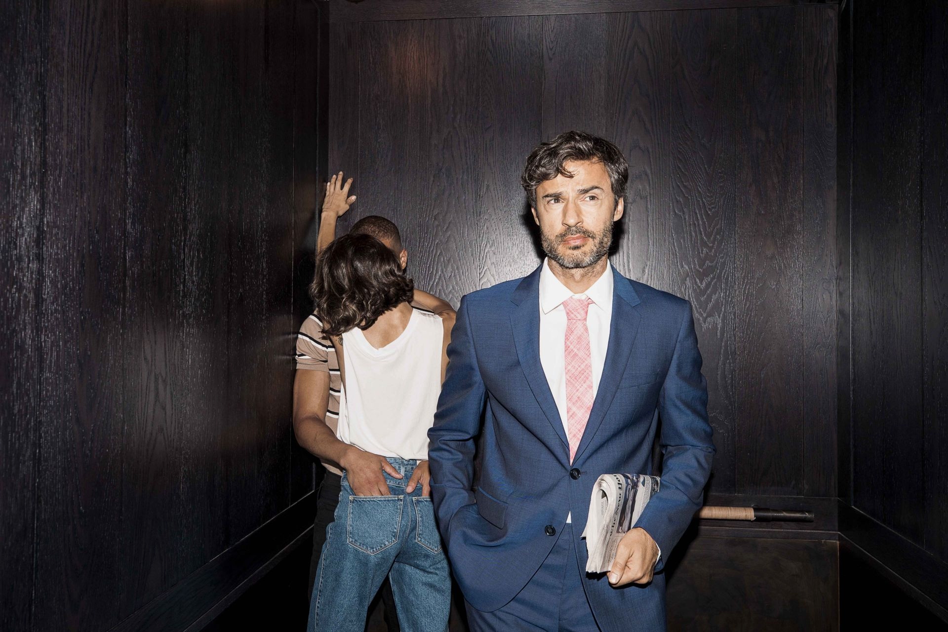 hip couple hugging with a suited man looking off in corner with his back to them